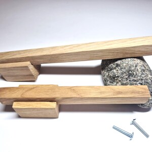 Könnte beinhalten: Zwei hellbraune Holzschrankgriffe mit rechteckigem Profil und glatter Oberfläche. Jeder Griff verfügt über einen zentralen Montageblock, der an einer langen Stange befestigt ist. Zwei silberne Metallschrauben sind für die Montage enthalten.