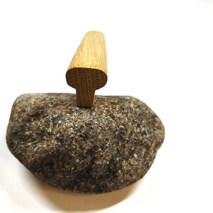 May include: A light brown wooden handle with a T-shaped profile is positioned vertically on top of a textured, multi-colored grey and brown natural stone. The wood grain is visible on the top surface of the handle, which has rounded edges.