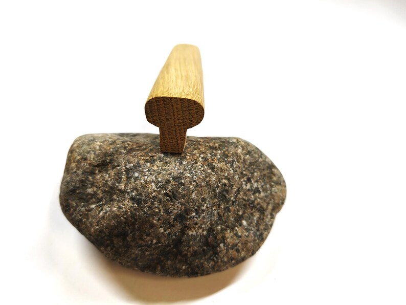 May include: A light brown wooden handle with a T-shaped profile is positioned vertically on top of a textured, multi-colored grey and brown natural stone. The wood grain is visible on the top surface of the handle, which has rounded edges.