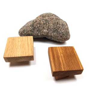 Könnte beinhalten: Zwei Holzknauf für Schränke, einer quadratisch und einer rechteckig, mit einer natürlichen Holzmaserung. Die Knäufe werden auf einer weißen Oberfläche neben einem grauen Stein gezeigt.