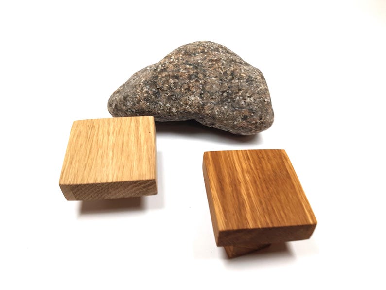 May include: Two wooden cabinet knobs, one square and one rectangular, with a natural wood grain finish. The knobs are displayed on a white surface next to a grey rock.