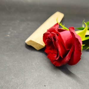 May include: A vibrant red rose with green leaves and stem, next to a small, light-colored wooden object. The rose is in full bloom, with its petals tightly curled. The background is a dark gray surface.