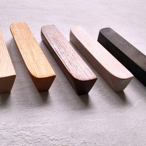 Puede incluir: Cinco tiradores de cajón de madera en diferentes manchas de madera. Los tiradores tienen todos la misma forma, con una parte superior redondeada y una parte inferior ligeramente puntiaguda. Los colores son madera natural, marrón claro, marrón oscuro, blanco y negro.