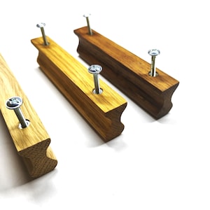 Puede incluir: Tres tiradores de cajón rectangulares de madera con perfil cóncavo, cada uno con dos tornillos metálicos plateados insertados por la parte superior. La veta de la madera es visible en las superficies marrón claro, medio y oscuro.
