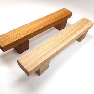 Puede incluir: Dos tiradores de cajón de madera, uno teñido de marrón oscuro y el otro dejado natural. Ambos tienen una forma rectangular con una sección central elevada y bordes cuadrados.