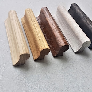 Pode incluir: Cinco puxadores de gaveta de madeira em diferentes acabamentos: madeira natural, madeira clara, madeira escura, branco e preto. Os puxadores têm todos a mesma forma, com uma parte superior curva e uma parte inferior plana.