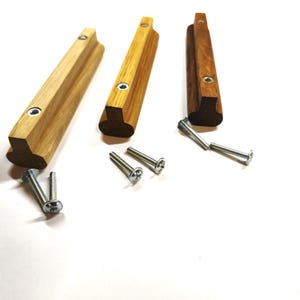 May include: Three wooden cabinet pulls in varying shades of light to dark brown, each featuring a T-shaped profile with two pre-drilled holes containing metal inserts. Six silver-colored metal screws are included for installation purposes.