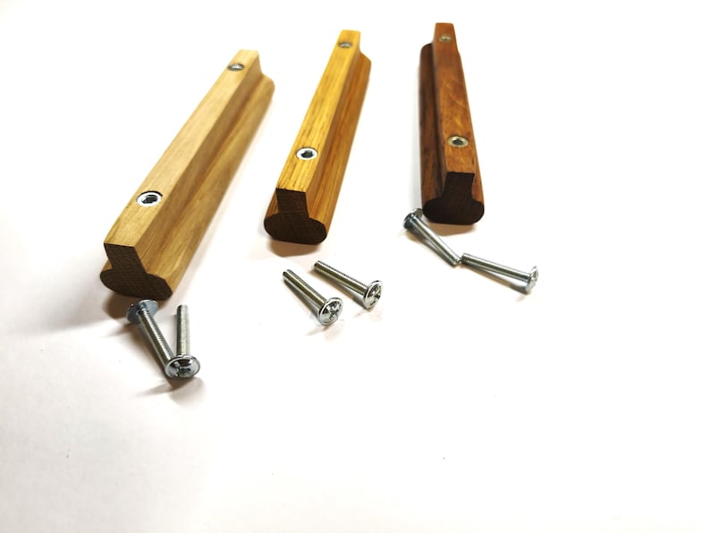 May include: Three wooden cabinet pulls in varying shades of light to dark brown, each featuring a T-shaped profile with two pre-drilled holes containing metal inserts. Six silver-colored metal screws are included for installation purposes.