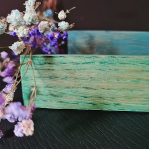 May include: A teal wooden box with a natural wood grain pattern. The box is empty and has a light blue painted interior. A bouquet of small white and purple flowers is in the foreground.