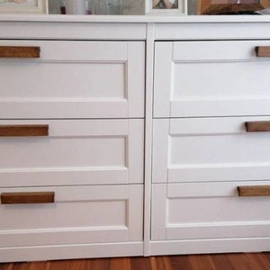 May include: A white dresser featuring six drawers arranged in two vertical columns. Each drawer is equipped with a rectangular, dark brown wooden handle. The drawer fronts have a recessed panel design, and the unit has a smooth, matte finish.