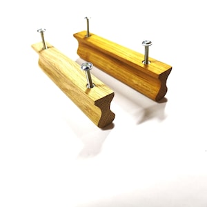May include: Two rectangular wooden drawer pulls with a light brown natural wood grain finish. Each handle features a concave profile along the front edge and is equipped with two silver metal screws protruding from the back for mounting purposes.