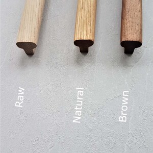 May include: Three wooden cabinet pulls with a T-shaped profile, displayed in three distinct wood tones labeled as Raw, Natural, and Brown. Each handle features a smooth, elongated top surface and a central mounting base, showcasing natural wood grain.