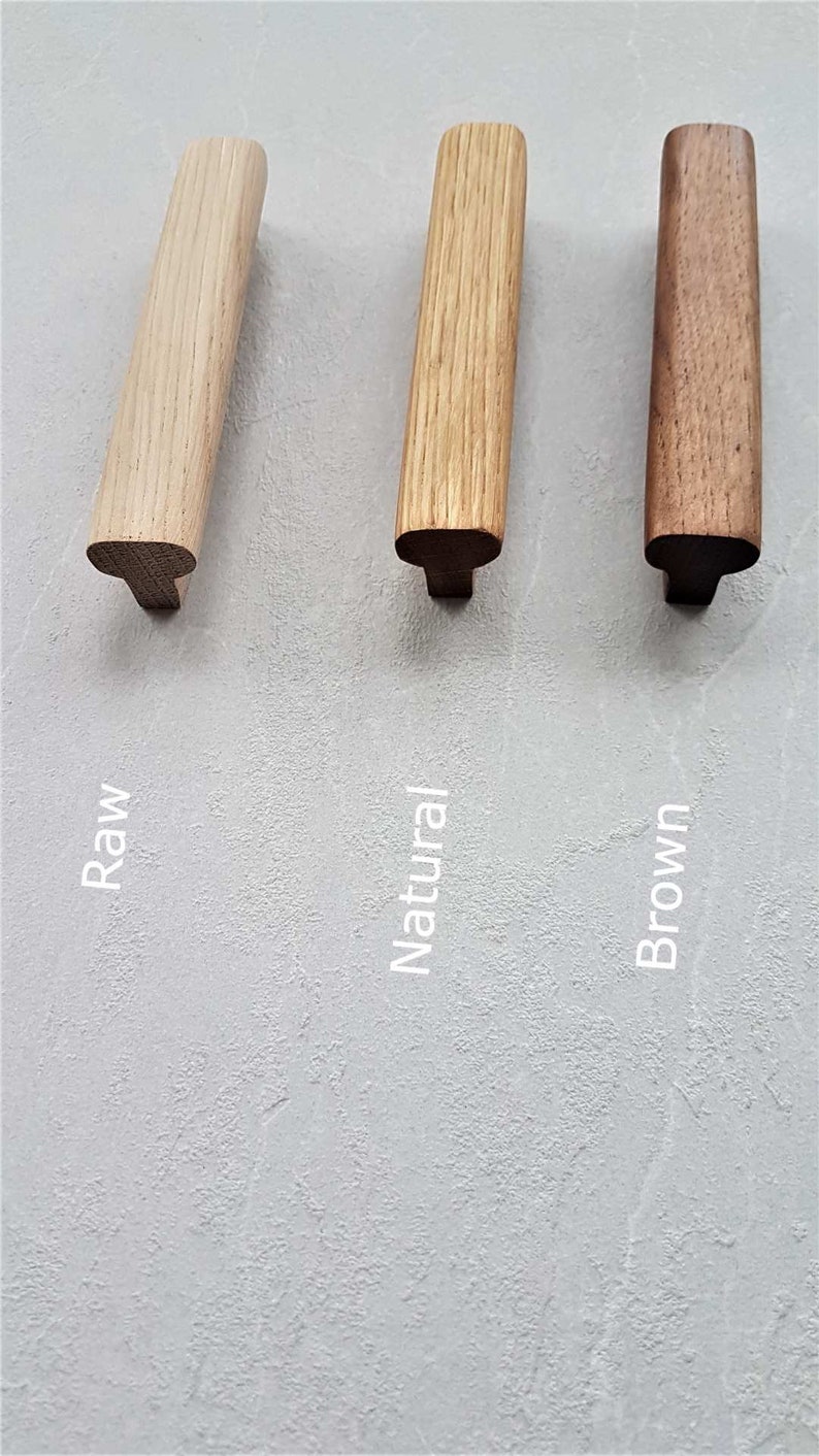 May include: Three wooden cabinet pulls with a T-shaped profile, displayed in three distinct wood tones labeled as Raw, Natural, and Brown. Each handle features a smooth, elongated top surface and a central mounting base, showcasing natural wood grain.