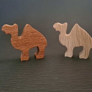 以下が含まれることがあります： 木製のラクダの置物2つ。1つは茶色、もう1つは薄い茶色。どちらも濃い灰色の表面の上に立っている。