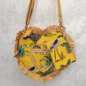 May include: A yellow heart-shaped crossbody bag with a matching small pouch. The bag and pouch feature a tropical bird and foliage print. The bag has a ruffled edge and a long, adjustable strap. The background is a textured, off-white wall.