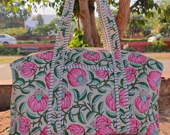May include: A large tote bag with a floral pattern. The bag features a cream-colored background with pink flowers and green leaves. The handles are white with a striped design. The bag is held up in front of a tree.
