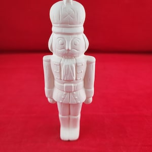 May include: A white, unpainted ceramic nutcracker figurine featuring a tall hat, a uniform with detailed buttons, a belt, and tall boots. The surface is matte and smooth, showing the molded facial features, including a mustache and eyes.