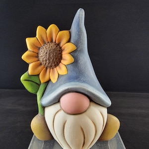 May include: A ceramic garden gnome figurine with a blue hat, a white beard, and a pink nose. The gnome holds a large sunflower with yellow petals and a brown center. The figurine is a decorative item.
