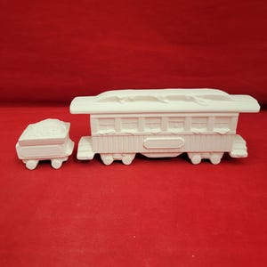May include: A white ceramic train set consisting of a passenger car and a smaller cargo car. The passenger car features rectangular windows and a roof, while the cargo car has a textured load. Both pieces are unpainted with a matte finish.
