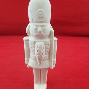 May include: A white, unpainted ceramic nutcracker figurine featuring a tall hat, a prominent mustache, a long beard, and a uniform with epaulets. The figure stands in an upright, rigid posture with a staff held vertically along the left side.