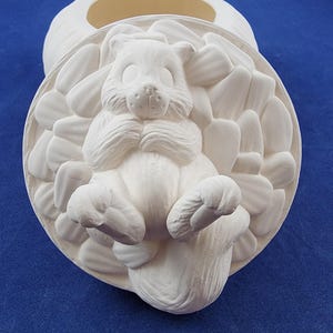 May include: White ceramic cookie jar with a squirrel design. The squirrel is sitting in a nest of leaves and has its paws up to its face. The lid of the jar is open and the inside is visible.