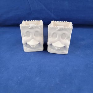 May include: Two white ceramic planters shaped like a cartoon face with a beak and eyes. The planters are on a blue surface.