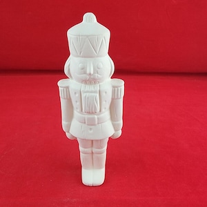 May include: A matte white ceramic nutcracker figurine featuring a tall hat, a uniform with epaulets, and a structured jacket. The surface is unpainted and smooth, showcasing the molded details of the soldier's facial features and attire.