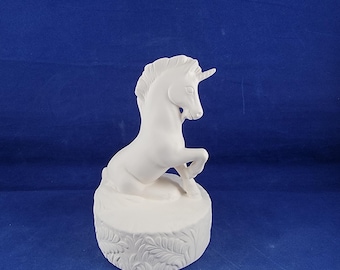 May include: White ceramic unicorn figurine with a horn, sitting on a round base with a leaf design.
