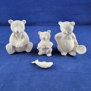 May include: A collection of unpainted ceramic bear figurines in various poses. One bear sits, another holds a fish, and a third holds a plate. A small fish figurine is also included. The items are on a blue background.