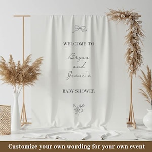 Op de afbeelding: Een witte stoffen achtergrond met de tekst "WELCOME TO Bryan and Jessie's BABY SHOWER" in zwart script. De achtergrond is versierd met een strik en bloemaccenten. Gedroogd pampasgras en bloemstukken omlijsten de achtergrond.