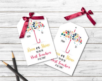 Op de afbeelding: Twee witte cadeau-etiketten met een kleurrijk paraplu-ontwerp en de tekst "Rain or Shine You Are The Best Teacher At All Time!" De etiketten zijn vastgebonden met rode linten.
