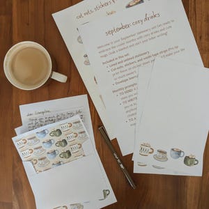 Puede incluir: Un juego de papelería con el tema de bebidas acogedoras, con papel rayado y sin rayar, forros de sobres y recortes. También hay un bolígrafo, una taza de café y una carta. El texto en el papel dice "septiembre: bebidas acogedoras."