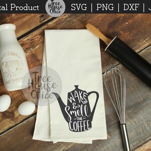 May include: A white tea towel with a black silhouette of a coffee pot and the text "WAKE & Smell -THE- COFFEE" printed on it. The tea towel is on a wooden surface with a rolling pin and a whisk.