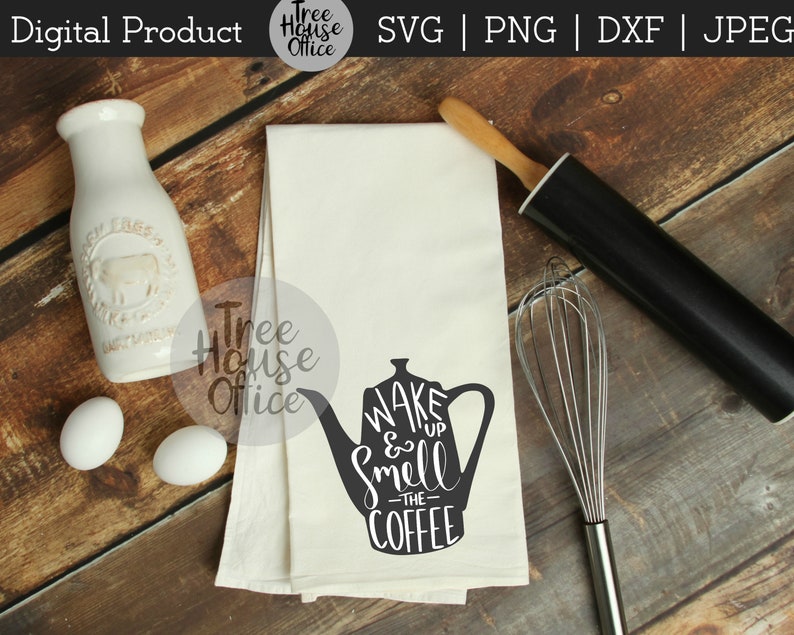 May include: A white tea towel with a black silhouette of a coffee pot and the text "WAKE & Smell -THE- COFFEE" printed on it. The tea towel is on a wooden surface with a rolling pin and a whisk.