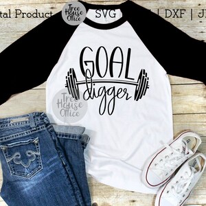 K&ouml;nnte beinhalten: Ein wei&szlig;es und schwarzes Baseball-T-Shirt mit dem Text "GOAL digger" und einer Hantelgrafik. Das Shirt liegt auf einer Holzoberfl&auml;che mit einem Paar wei&szlig;en Turnschuhen und einer blauen Jeans.