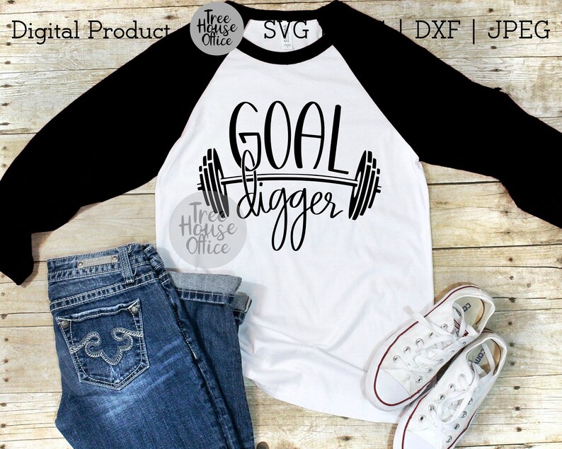 K&ouml;nnte beinhalten: Ein wei&szlig;es und schwarzes Baseball-T-Shirt mit dem Text "GOAL digger" und einer Hantelgrafik. Das Shirt liegt auf einer Holzoberfl&auml;che mit einem Paar wei&szlig;en Turnschuhen und einer blauen Jeans.