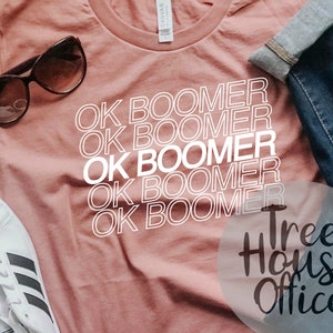 May include: A pink t-shirt with the phrase "OK Boomer" repeated five times in white text. The shirt is laying on a table with a pair of sunglasses and a pair of jeans. The text "Tree House Office" is visible in the lower right corner of the image.