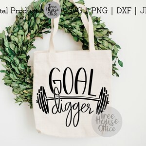 K&ouml;nnte beinhalten: Eine wei&szlig;e Einkaufstasche mit schwarzem Text, der "GOAL digger" lautet, mit einer Langhantel und Gewichtsscheiben auf beiden Seiten des Textes. Die Einkaufstasche liegt auf einem gr&uuml;nen Kranz.