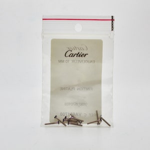 May include: A small clear plastic bag with a white label that reads 'Cartier' in black text. The label also has the text 'EN101JUEV.101MM' in black text. The bag contains a small quantity of silver metal pins.