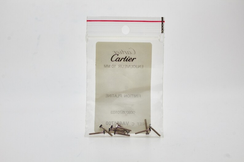 May include: A small clear plastic bag with a white label that reads 'Cartier' in black text. The label also has the text 'EN101JUEV.101MM' in black text. The bag contains a small quantity of silver metal pins.
