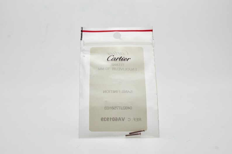 May include: A small clear plastic bag with a red zipper seal. The bag contains two small silver metal pins. The bag has a white label with the text 'Cartier', 'ENVOLUTION 10 MM', 'SANS FINITION', '04297728103', 'REF: C.VAG01829'. 