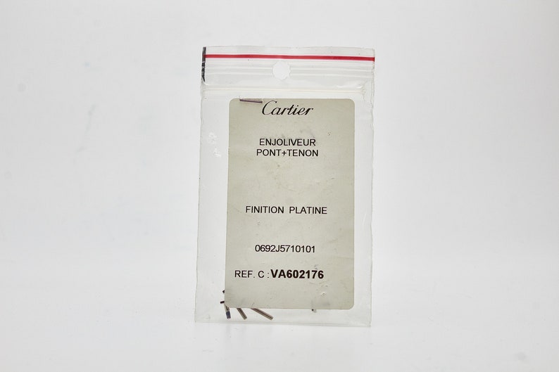 May include: A white card with black text in a clear plastic bag. The text reads: 'Cartier', 'Enjoliveur Pont + Tenon', 'Finition Platine', '0692J5710101', 'REF. C: VA602176'