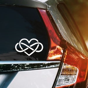 May include: White vinyl car decal featuring a heart intertwined with an infinity symbol. The decal is affixed to the rear window of a silver car, with a sunny road in the background. The design is simple and elegant.