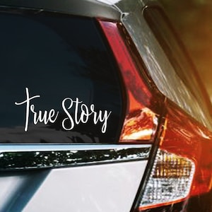 May include: A white vinyl car decal with the words "True Story" in a cursive font is affixed to the rear window of a silver car. The car's taillight is visible, and the background is blurred.