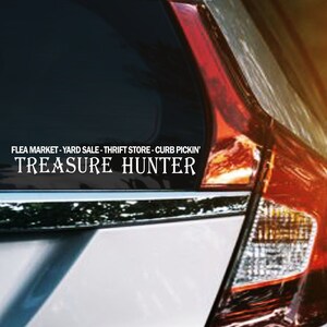 May include: A silver car with a sticker on the rear window that reads "FLEA MARKET - YARD SALE - THRIFT STORE - CURB PICKIN' TREASURE HUNTER". The car's taillight is red and orange, and the side mirror is visible.