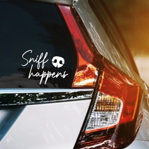 May include: A white vinyl car decal with the text "Sniff happens" in a cursive font, accompanied by a graphic of a dog's nose. The decal is affixed to the rear window of a silver car, with the taillight and side mirror visible.