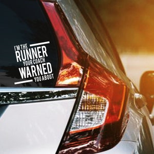 May include: A white vinyl decal on a car's rear window. The decal reads "I'M THE RUNNER YOUR COACH WARNED YOU ABOUT" in a bold, modern font. The car is silver with a red taillight, parked on a road with a blurred background.