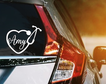 May include: White vinyl decal of a heart with a stethoscope design and the name "Amy" in cursive. The decal is affixed to the rear window of a car. The car is silver with a red tail light.