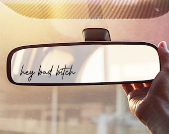 Puede incluir: Espejo retrovisor de coche con la frase "hey bad bitch" en cursiva. Una mano sostiene el espejo, con el interior del coche y el parabrisas visibles. La imagen tiene un tono cálido y soleado.