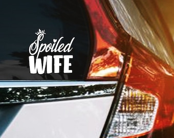 Puede incluir: Una calcomanía blanca para coche con las palabras "Spoiled Wife" en una fuente cursiva, coronada con una corona. La calcomanía está adherida a la ventana trasera de un coche plateado, con un fondo borroso.
